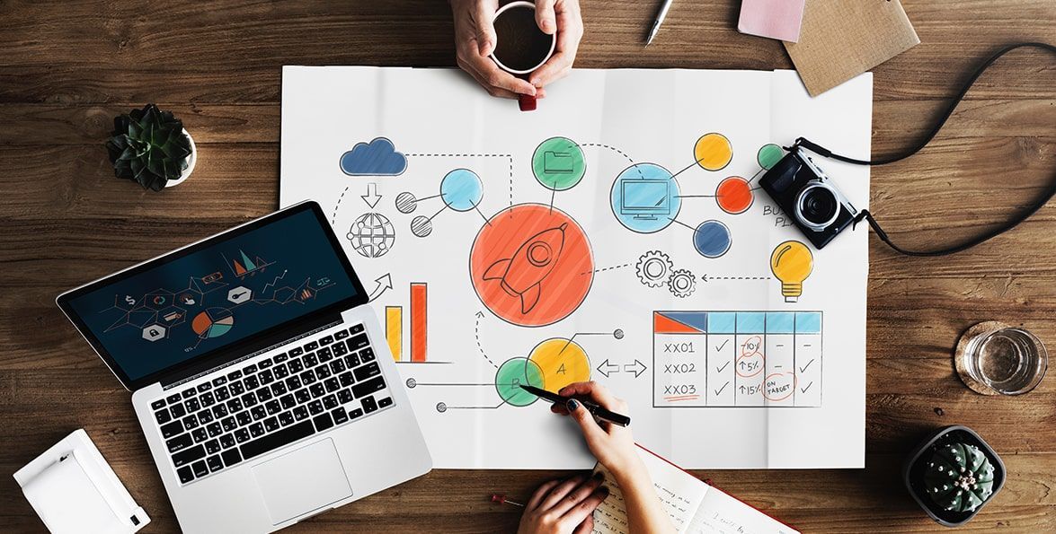 Vista cenital de una mesa de trabajo con una computadora portátil y un afiche de planificación con diagramas, gráficos y un cohete dibujado