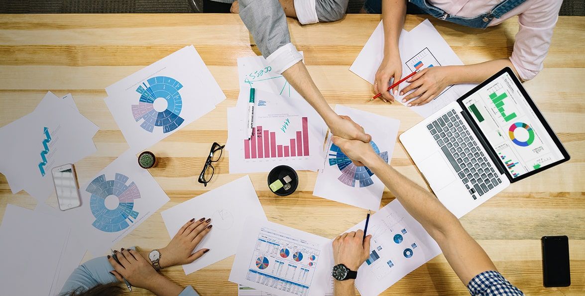 Manos de un equipo que se estrechan sobre una mesa con informes, gráficos de barras y circulares y una tablet