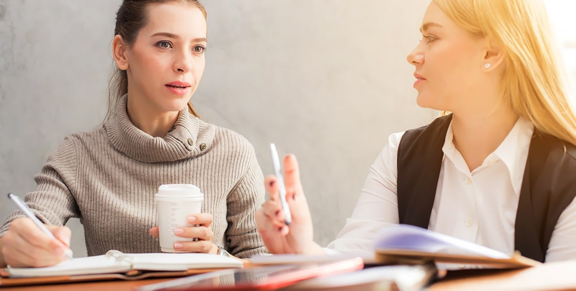 Dos mujeres conversan sentadas a una mesa con cuadernos y un vaso de café