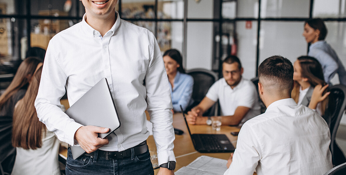Hombre sonriente con una carpeta en un espacio de trabajo compartido, con personas reunidas alrededor de una mesa