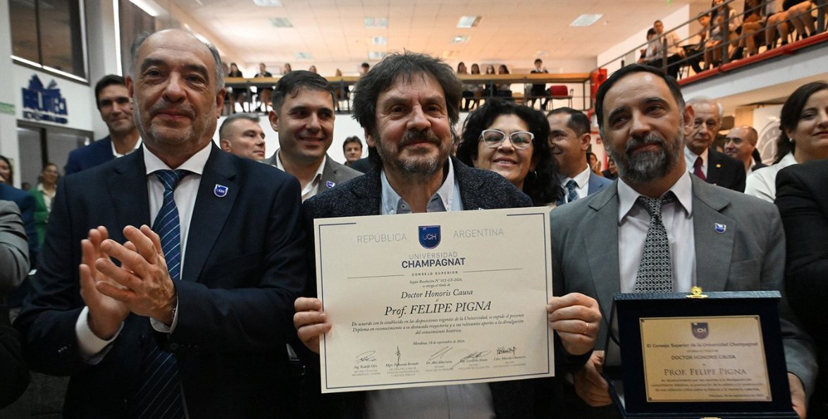 Felipe Pigna sostiene el diploma de Doctor Honoris Causa junto al rector Marcelo Chamorro y autoridades, que aplauden en la biblioteca de la Universidad.
