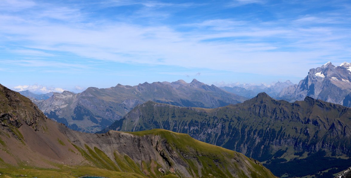 Paisaje de alta montaña con cordones rocosos, laderas verdes y cielo azul.