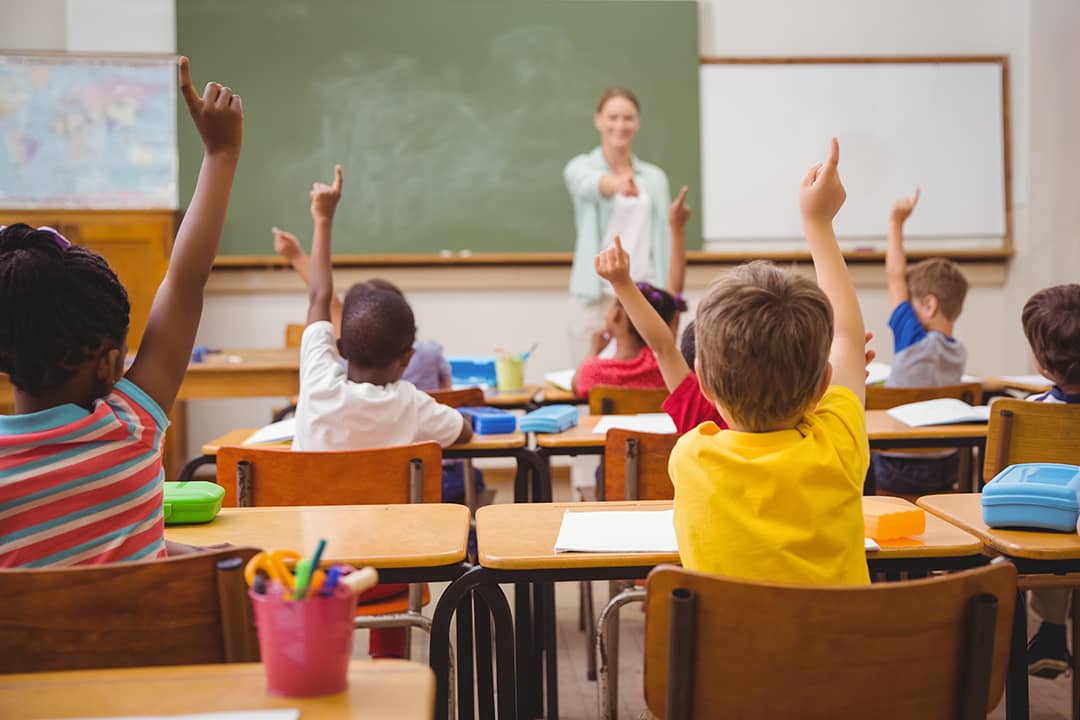 Aula de primaria: niñas y niños levantan la mano frente a una docente junto al pizarrón